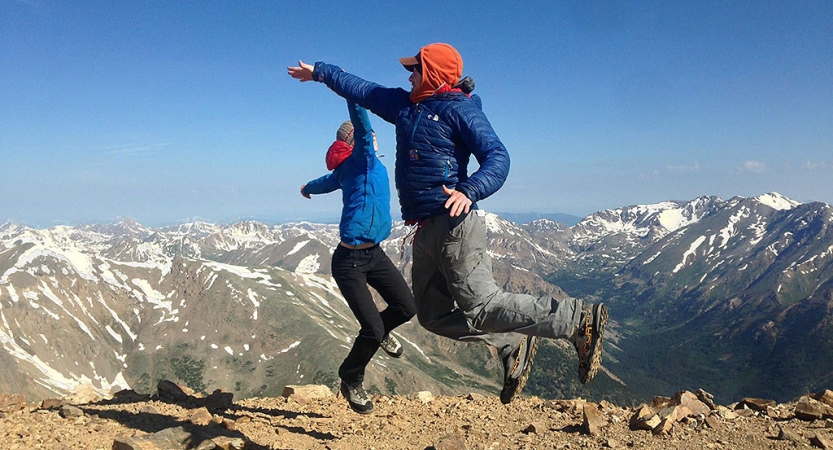 Two people jump in the air and high five with a mountainous landscape in the background
